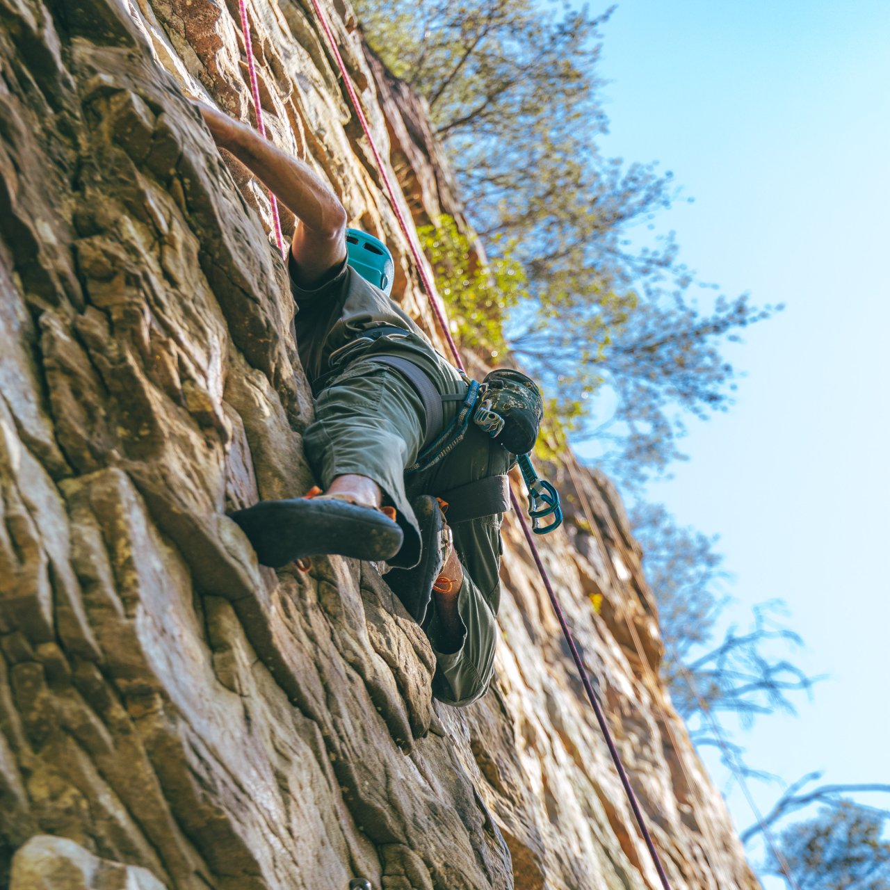 A climber with a blue helmet scales a rugged rock face, surrounded by trees under a clear blue sky. The scene conveys adventure and determination.