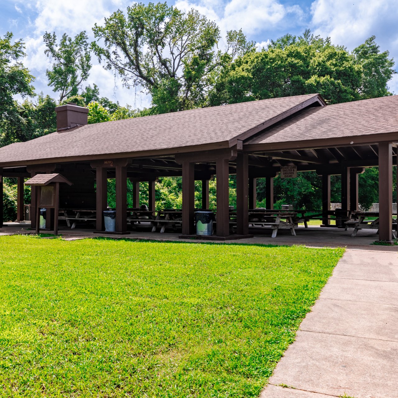A spacious wooden picnic pavilion under a partly cloudy sky, surrounded by lush green grass and trees. The atmosphere is serene and inviting.