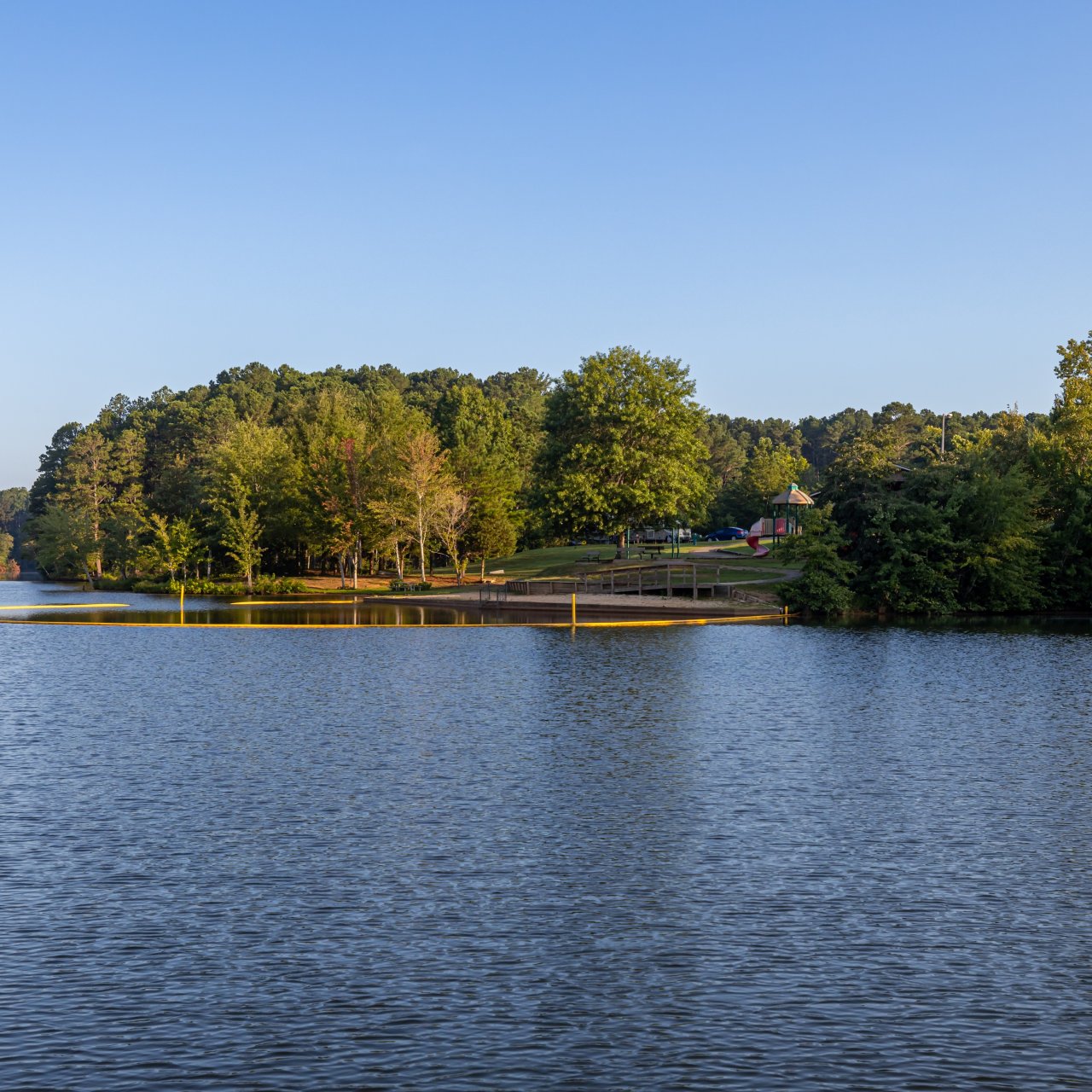 Serene lake scene with calm water reflecting trees and a small, sandy beach in the background. Green forest surrounds the area under a clear blue sky.
