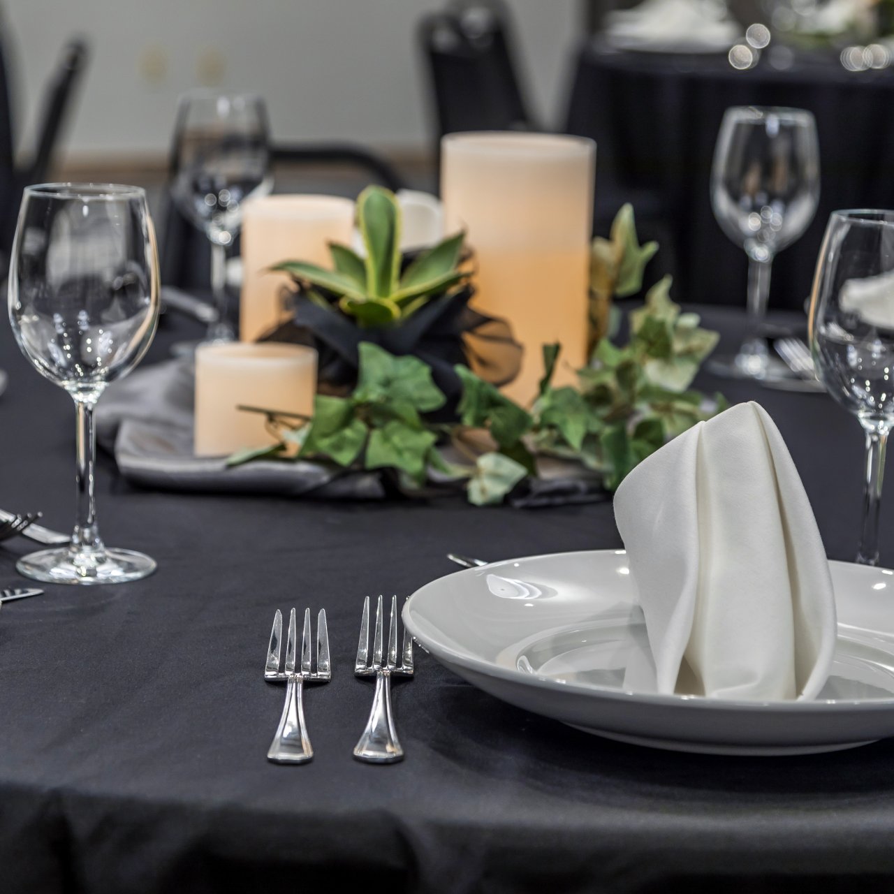Elegantly set dining table with black cloth, white plates, folded napkins, wine glasses, and silverware. Central candles and greenery add a warm, inviting ambiance.