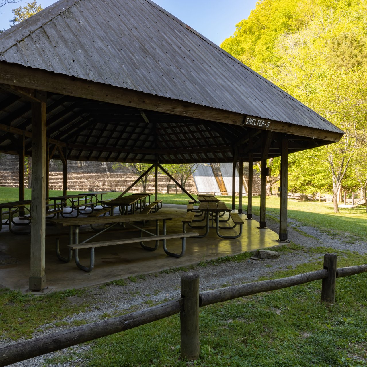 A wooden park shelter with picnic tables in a sunlit grassy area, surrounded by lush green trees. Relaxed, inviting outdoor setting.