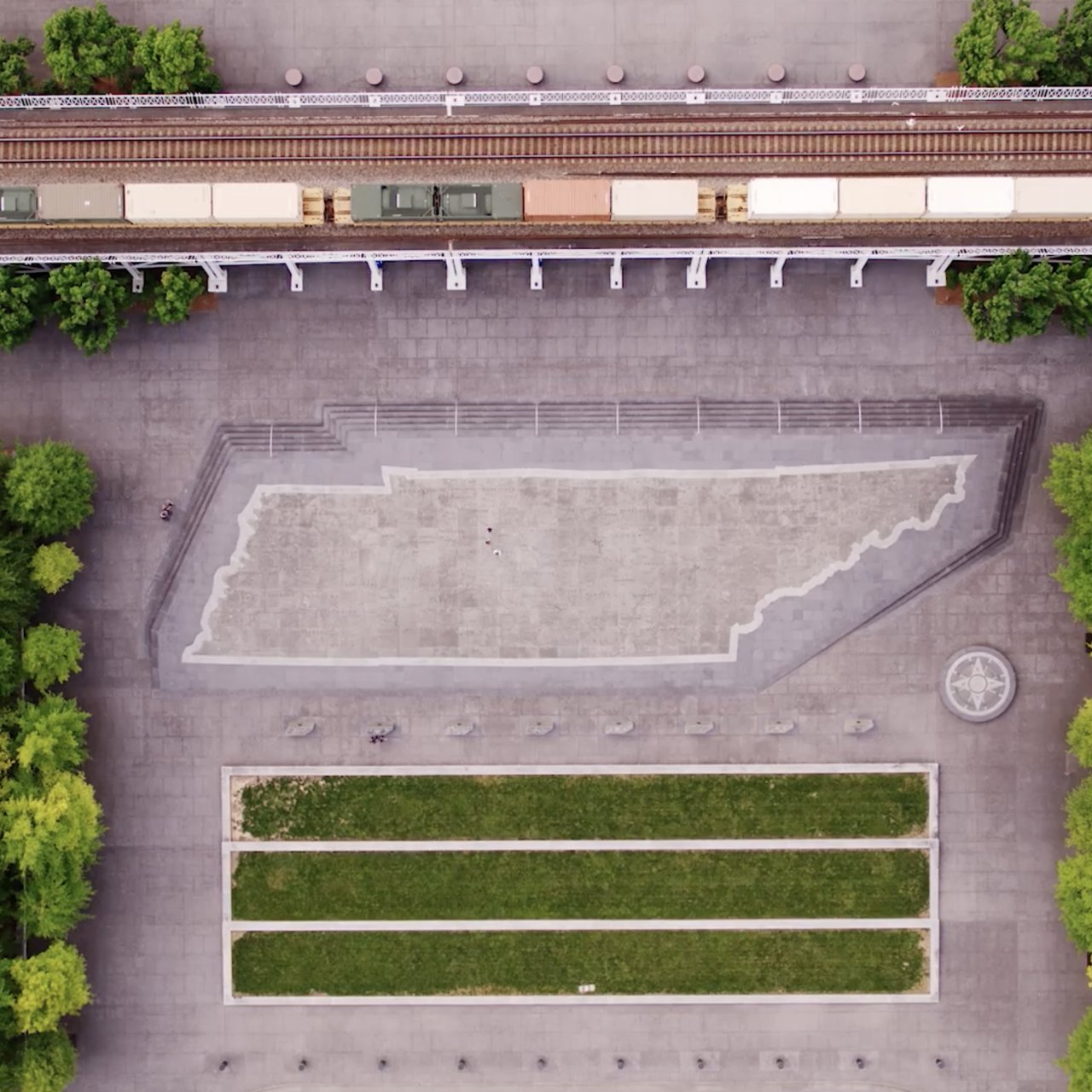 Aerial view of paved plaza featuring a large, outlined Tennessee shape. Train tracks run above, bordered by lush green trees lining the pathways.