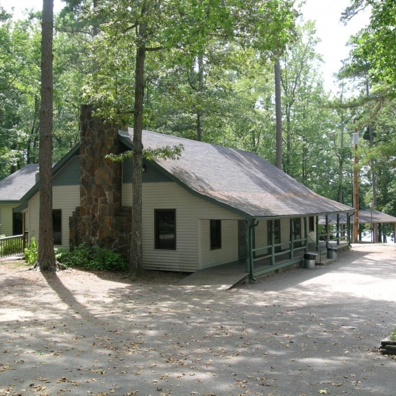 A rustic cabin with a stone chimney nestled among tall trees. The setting is serene, with sunlight dappling the roof and leafy shadows on the ground.