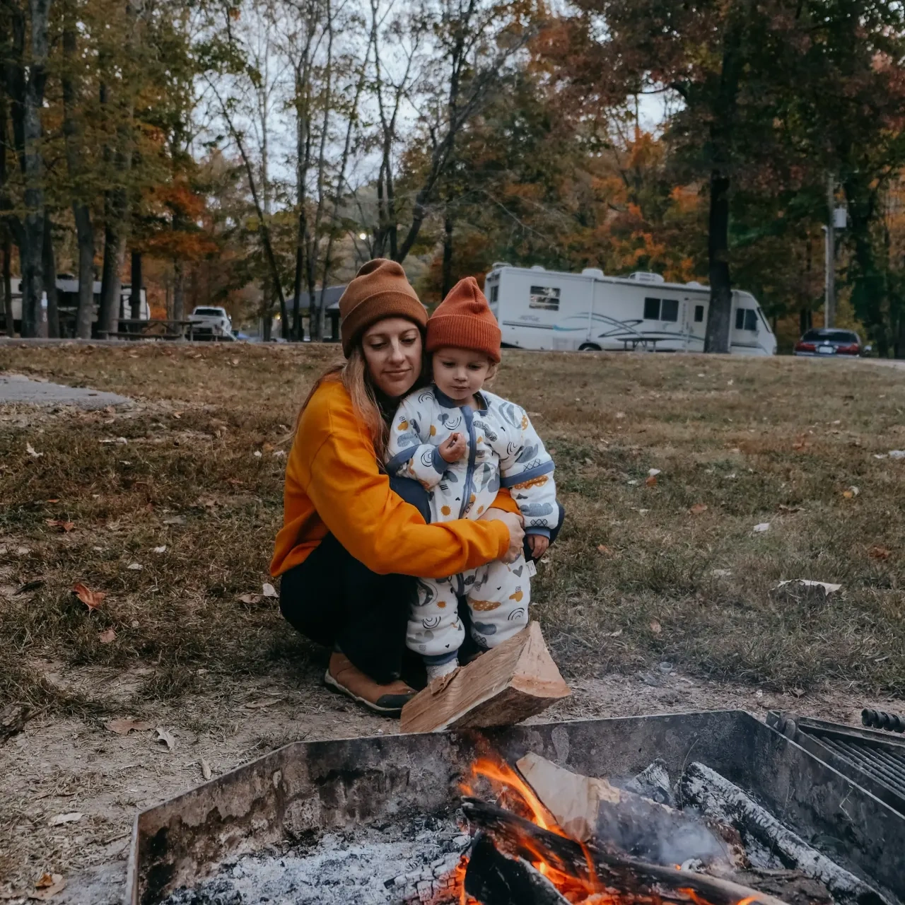 Woman and child in hats by a campfire, surrounded by autumn trees.