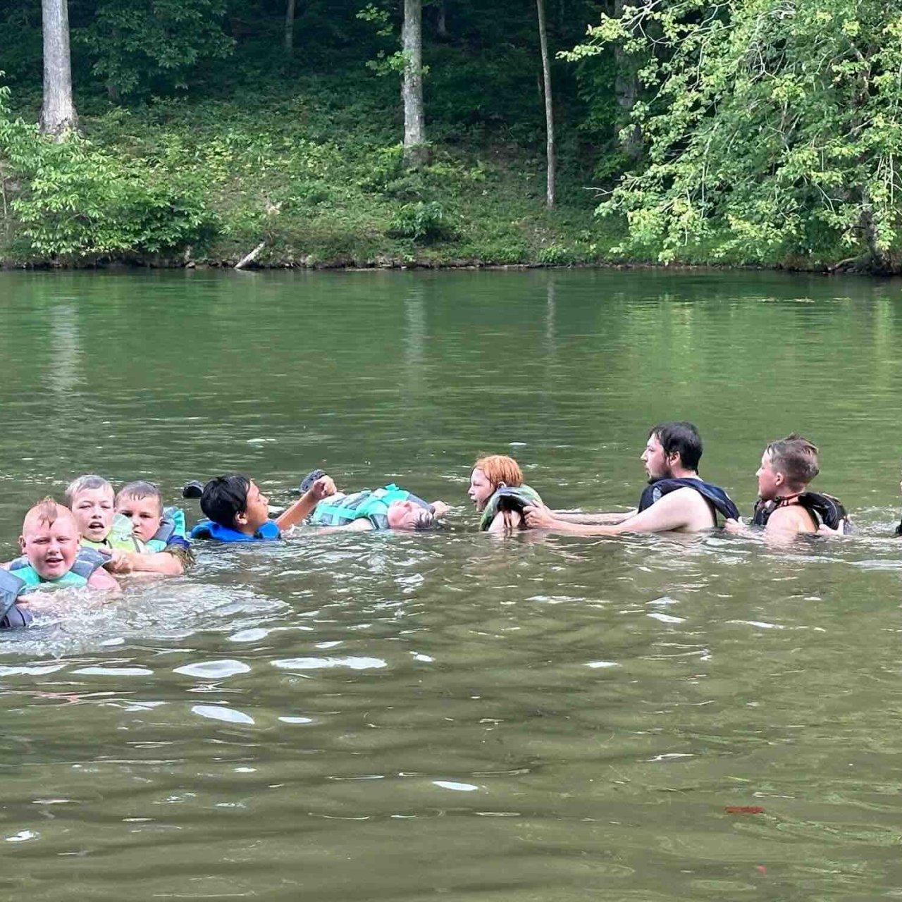 group of kids swimming in a line, holding on to each other with life jackets on