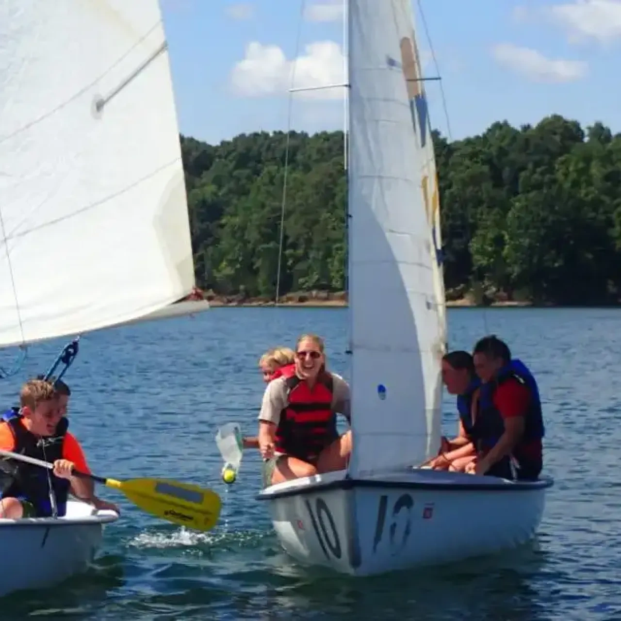 Two small sailboats with people in life vests on a sunny lake.