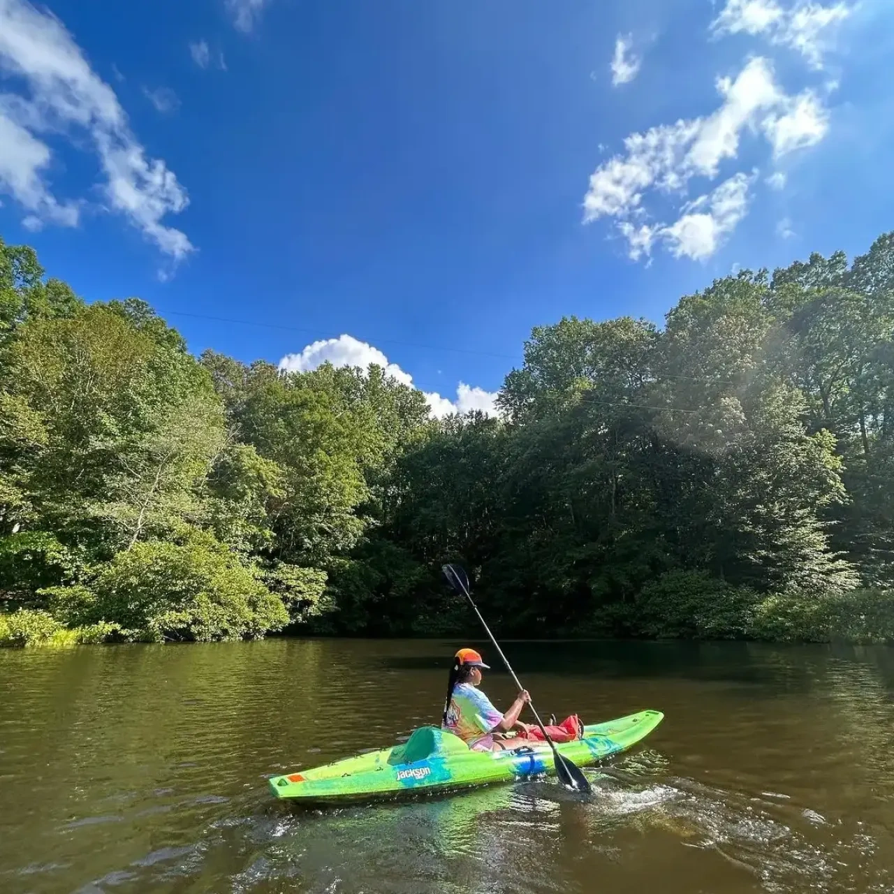 A person paddles a green kayak on a river under a bright blue sky.
