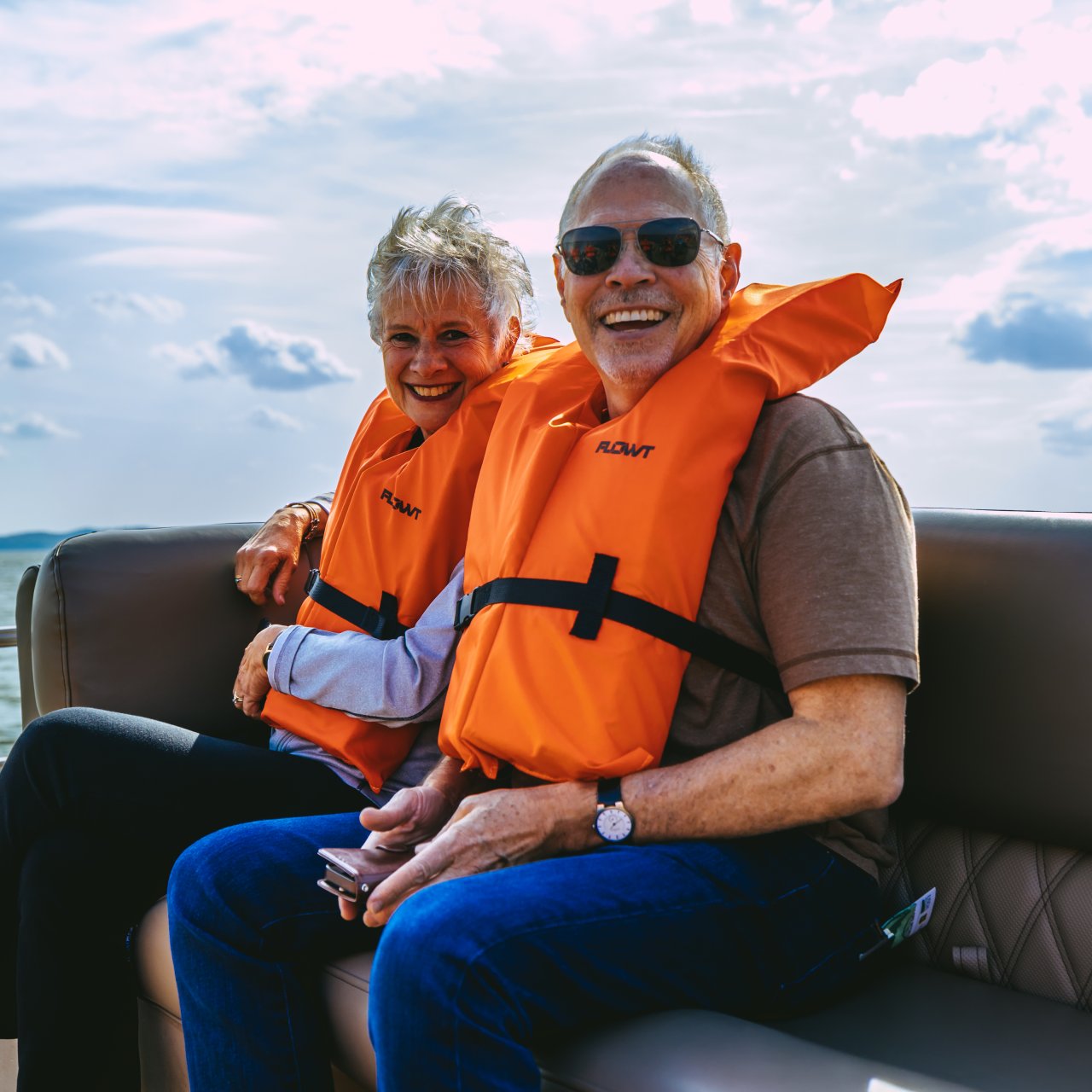 Elderly couple wearing orange life jackets sit smiling on a boat, with a calm lake and cloudy sky in the background. They appear joyful and relaxed.
