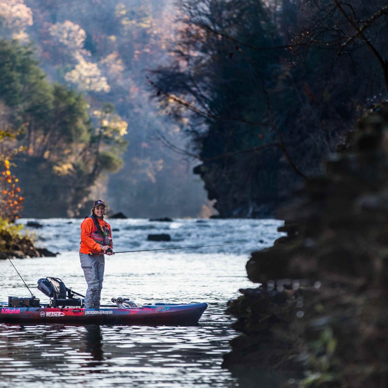 A person in an orange jacket stands on a kayak, fishing on a calm river surrounded by lush, autumn-colored trees, conveying peace and focus.