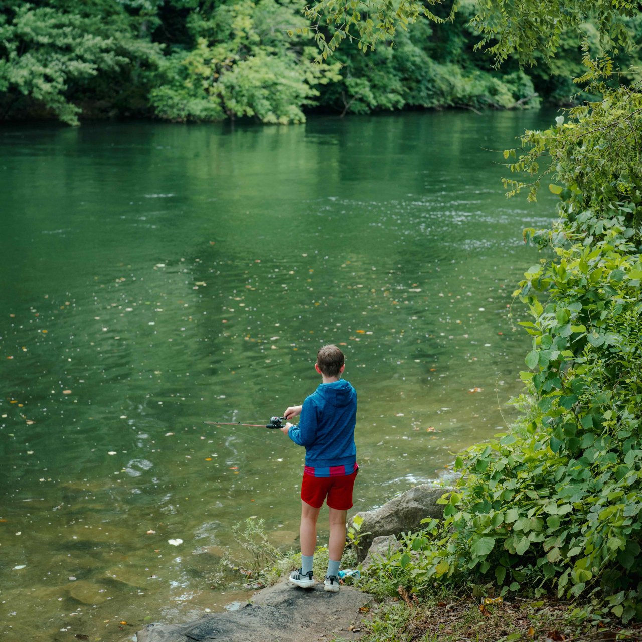 boy wearing shorts and a hoodie fishing in a river next to some trees