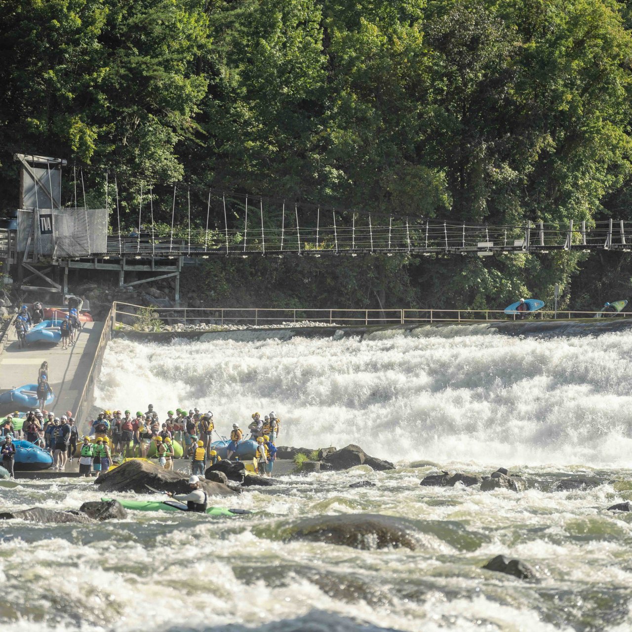 Area for white water rafting where there is a slope for rafts to go into water with tons of people and a bridge in the background