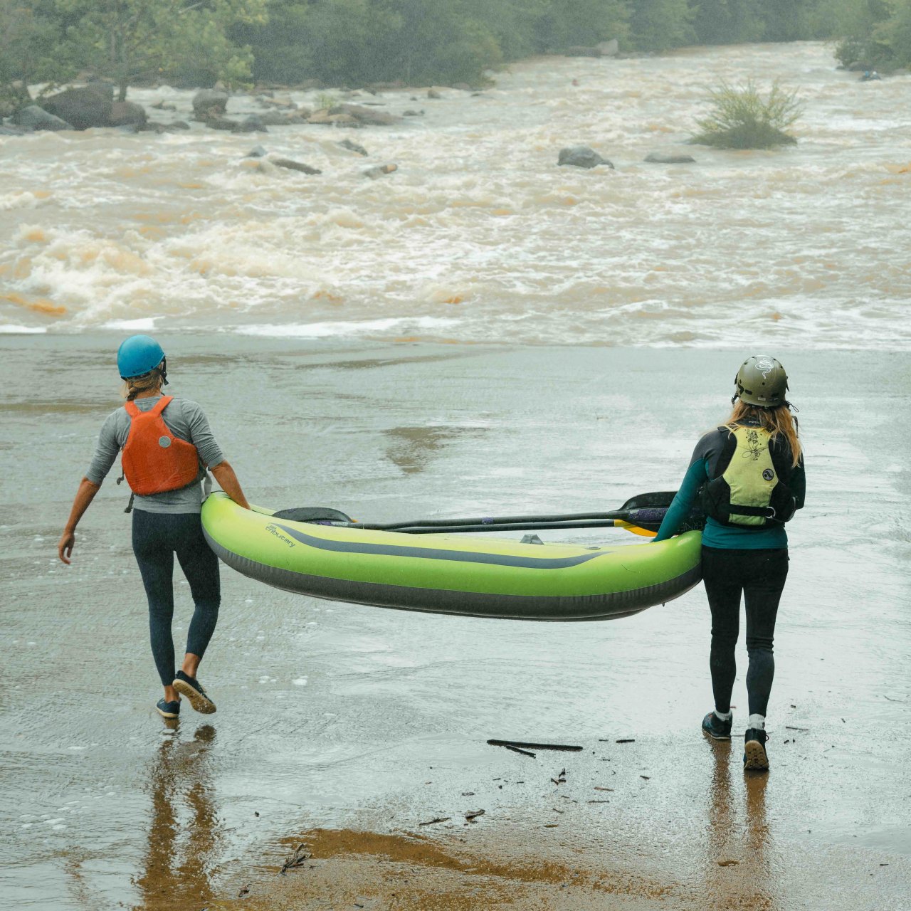 two women wearing helmets and vests on opposite sides of a raft, carrying it into the water on a cloudy day