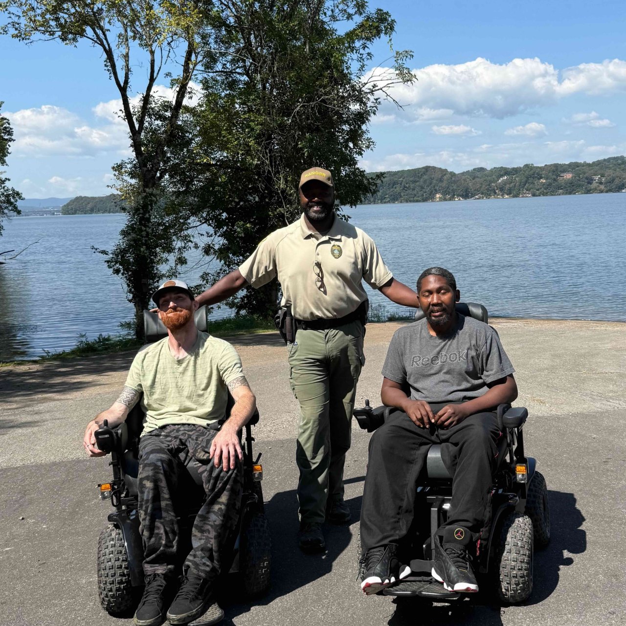 Two men in wheelchairs and a park ranger standing by a lake.