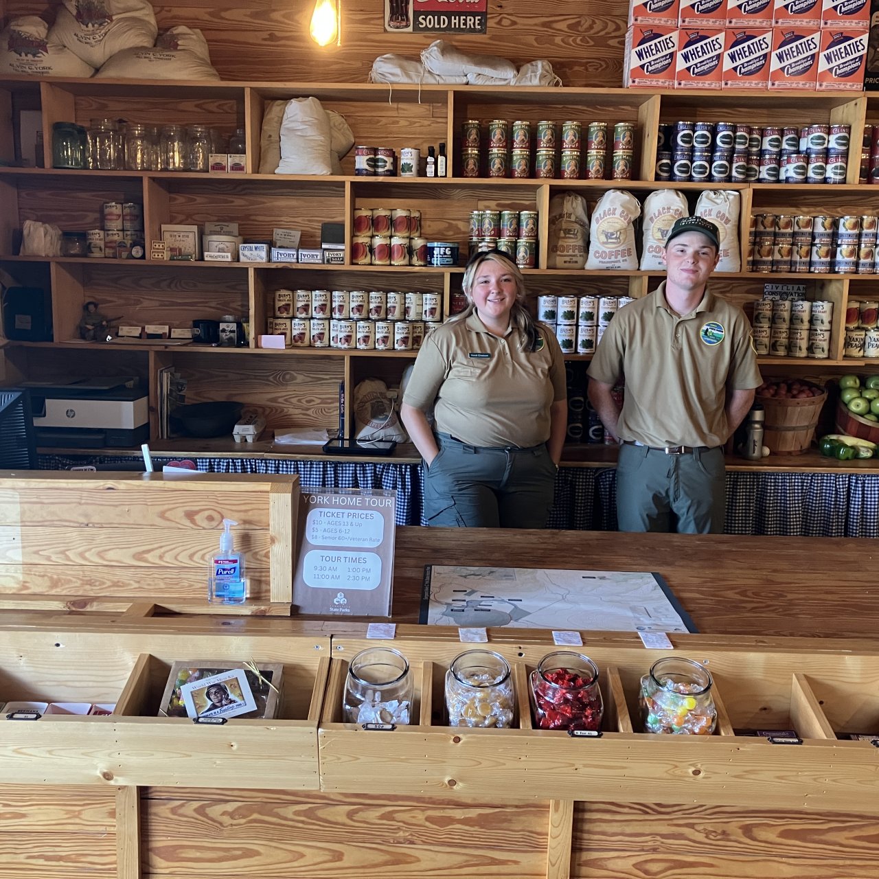 Two employees stand behind a rustic wooden counter in a general store.
