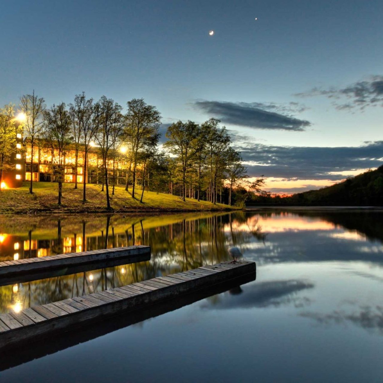 Twilight scene with lit trees reflecting on a calm lake, dock in foreground.