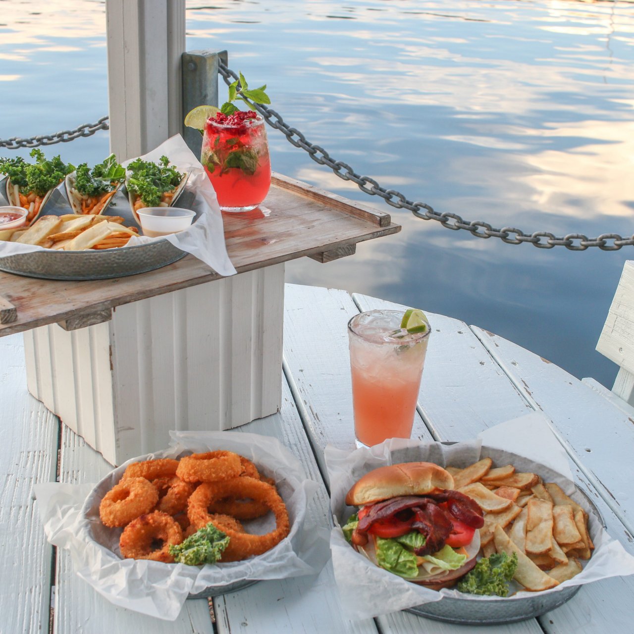 Outdoor meal on a dock with a burger, fries, onion rings, and drinks overlooking water.