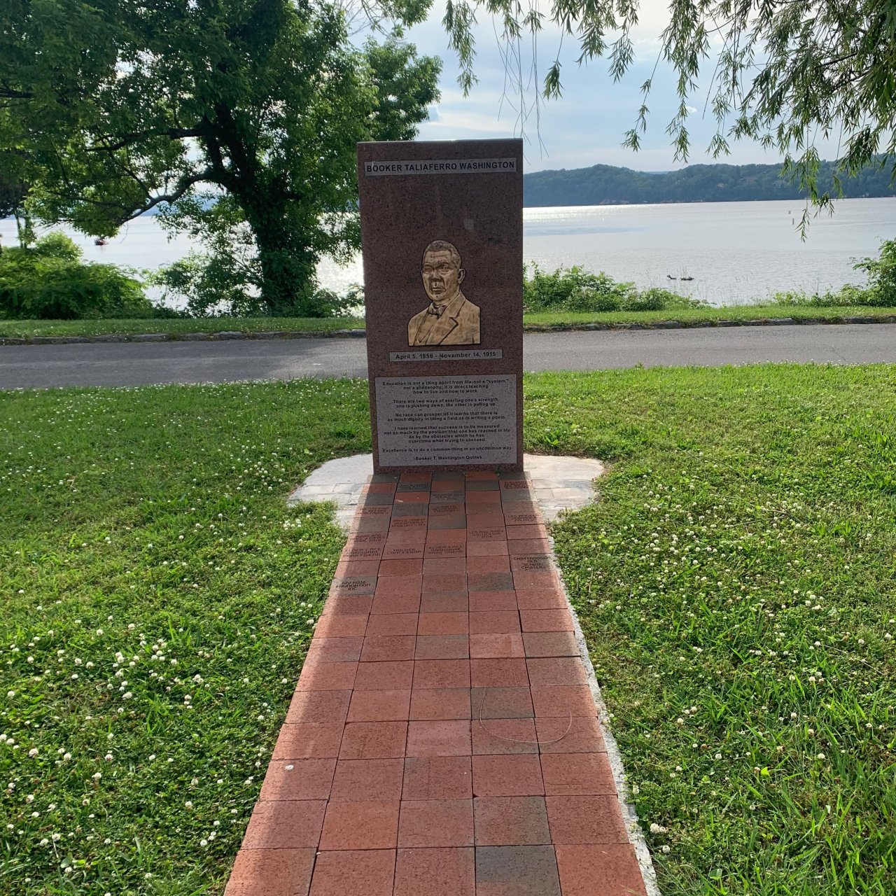 Bust monument on a brick path by a lake, surrounded by grass and trees.