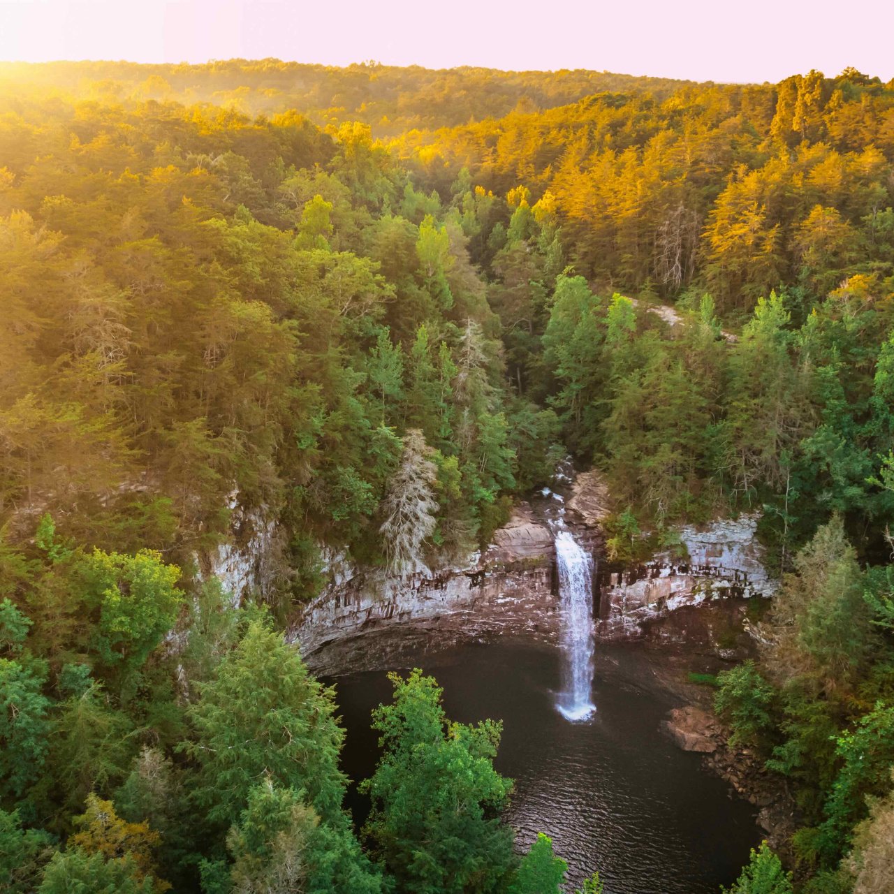 Sunrise over a forest with a waterfall flowing into a serene pond.