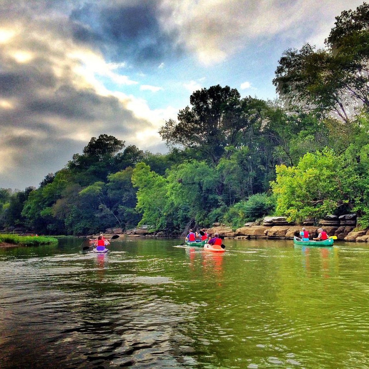 Kayakers on a river with lush green trees under a cloudy sky.