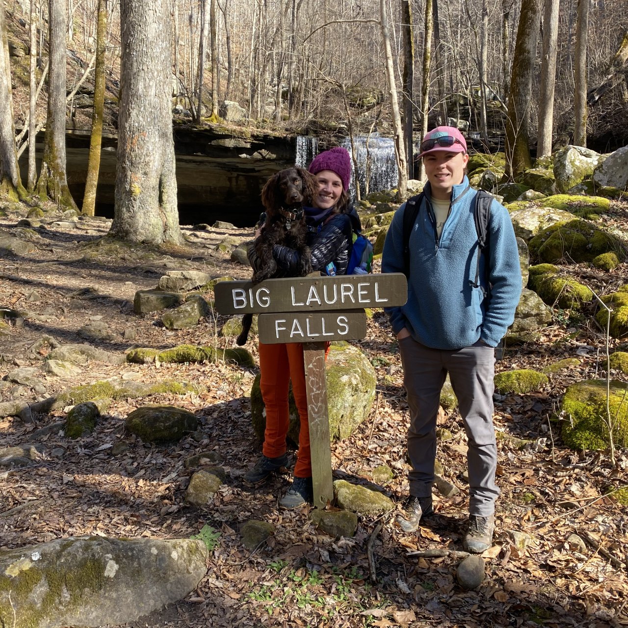 Couple with dog by sign for Big Laurel Falls in a wooded area.