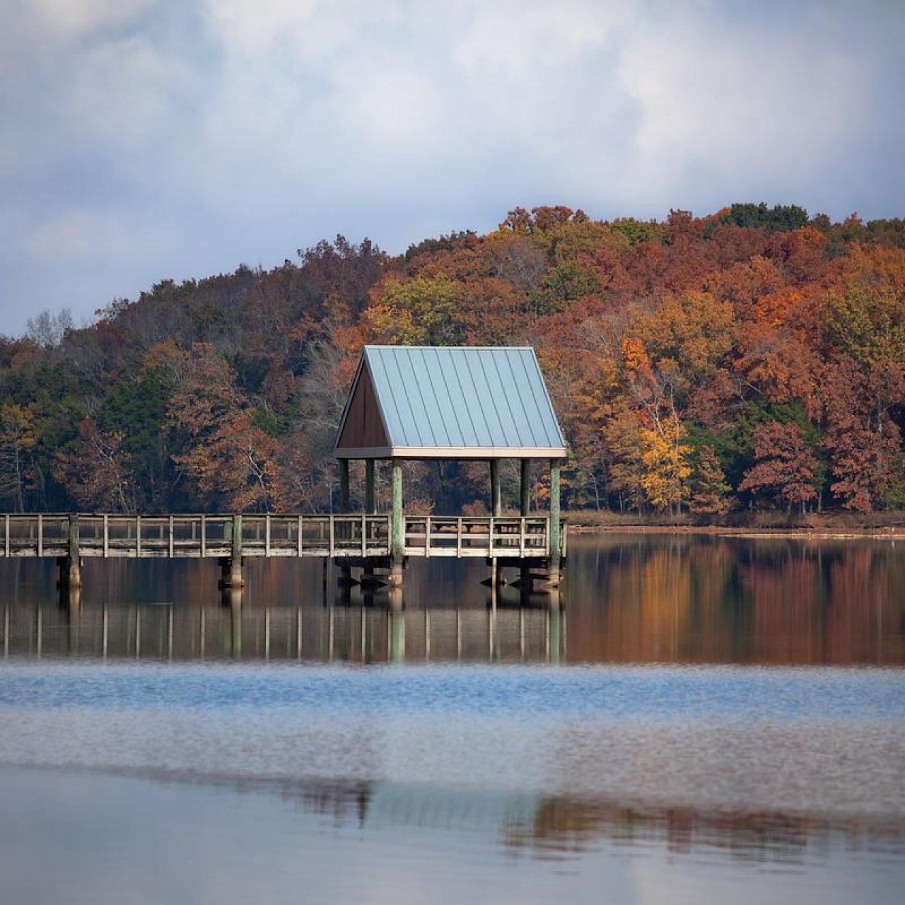 Small gazebo on a pier overlooking a tranquil lake with autumn trees.