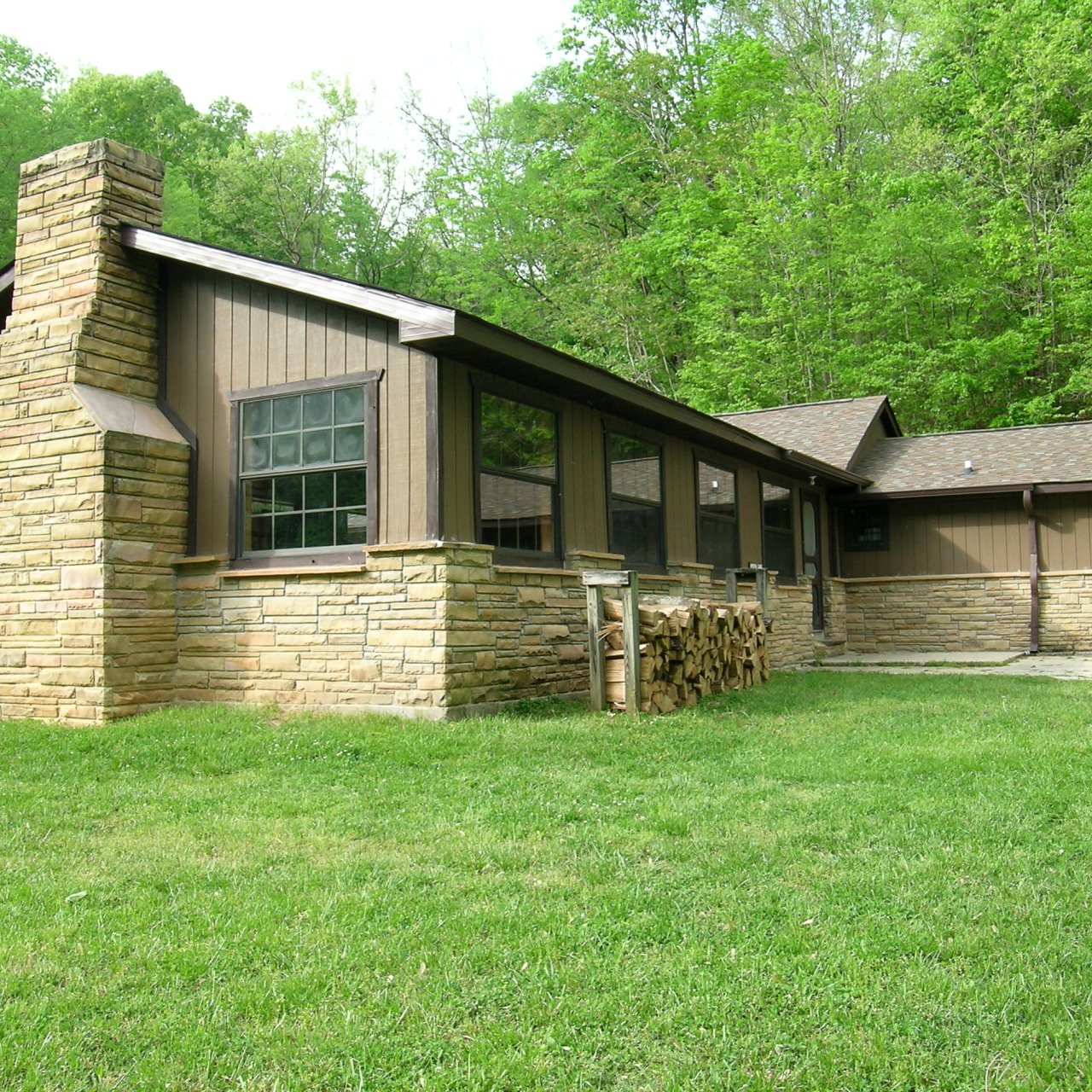 Cabin with stone chimney surrounded by lush green grass and trees.