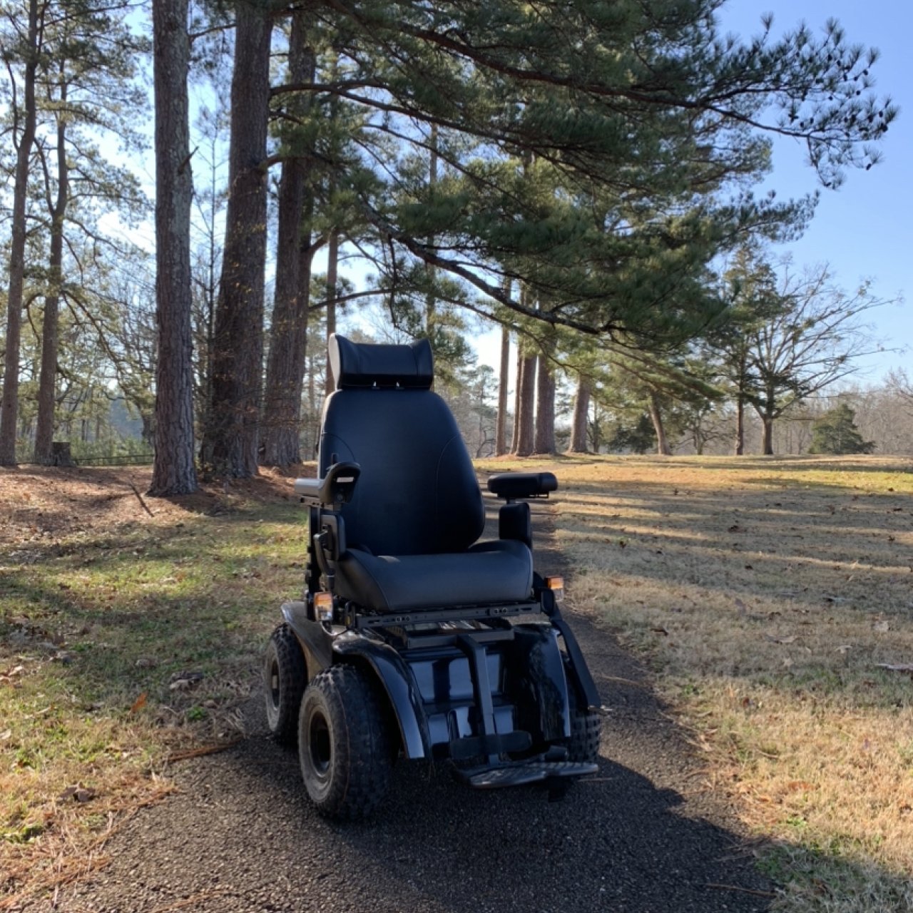 An all-terrain wheelchair sits upon a paved path with tall pine trees behind it.