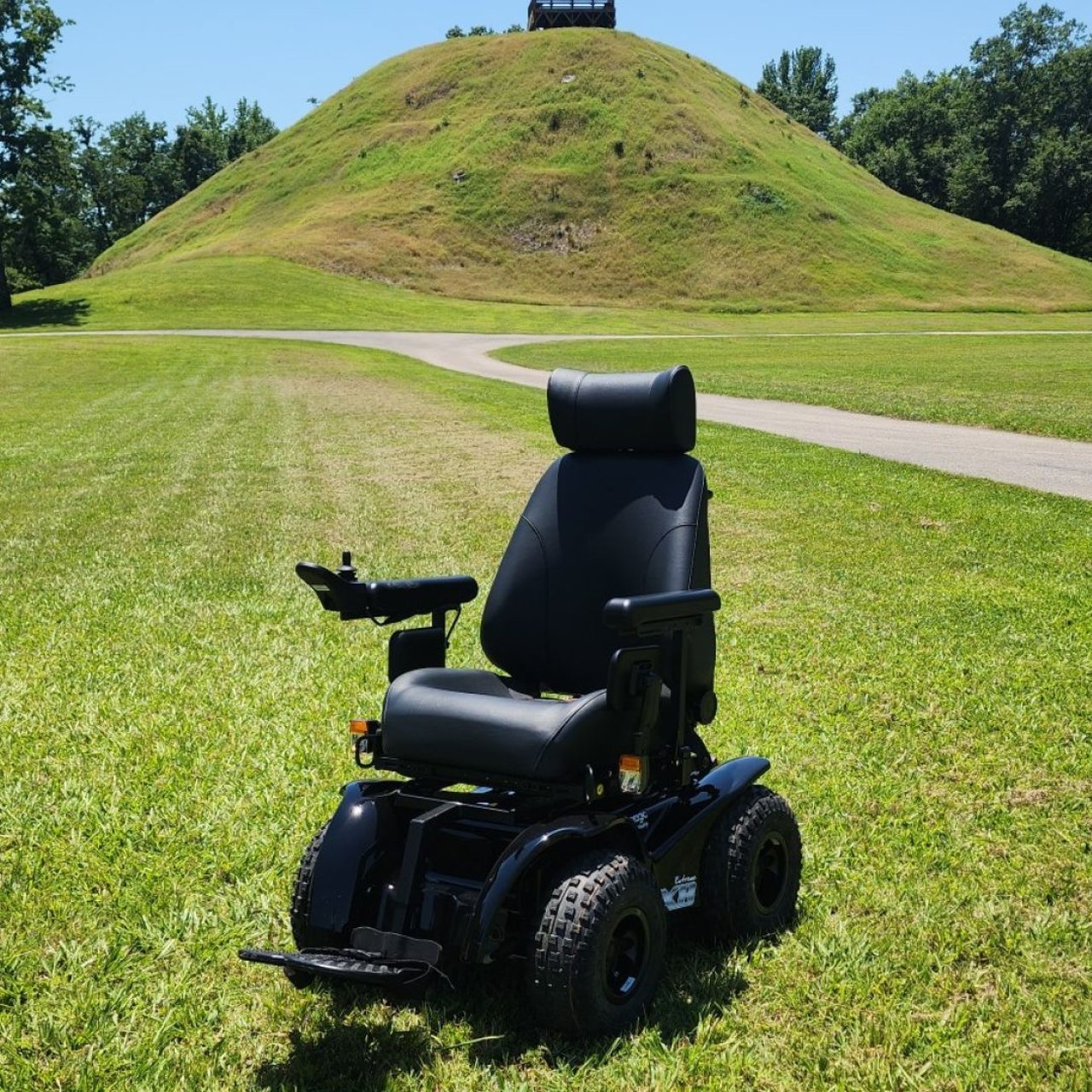 All terrain wheelchair in the grass with a giant mound behind it.