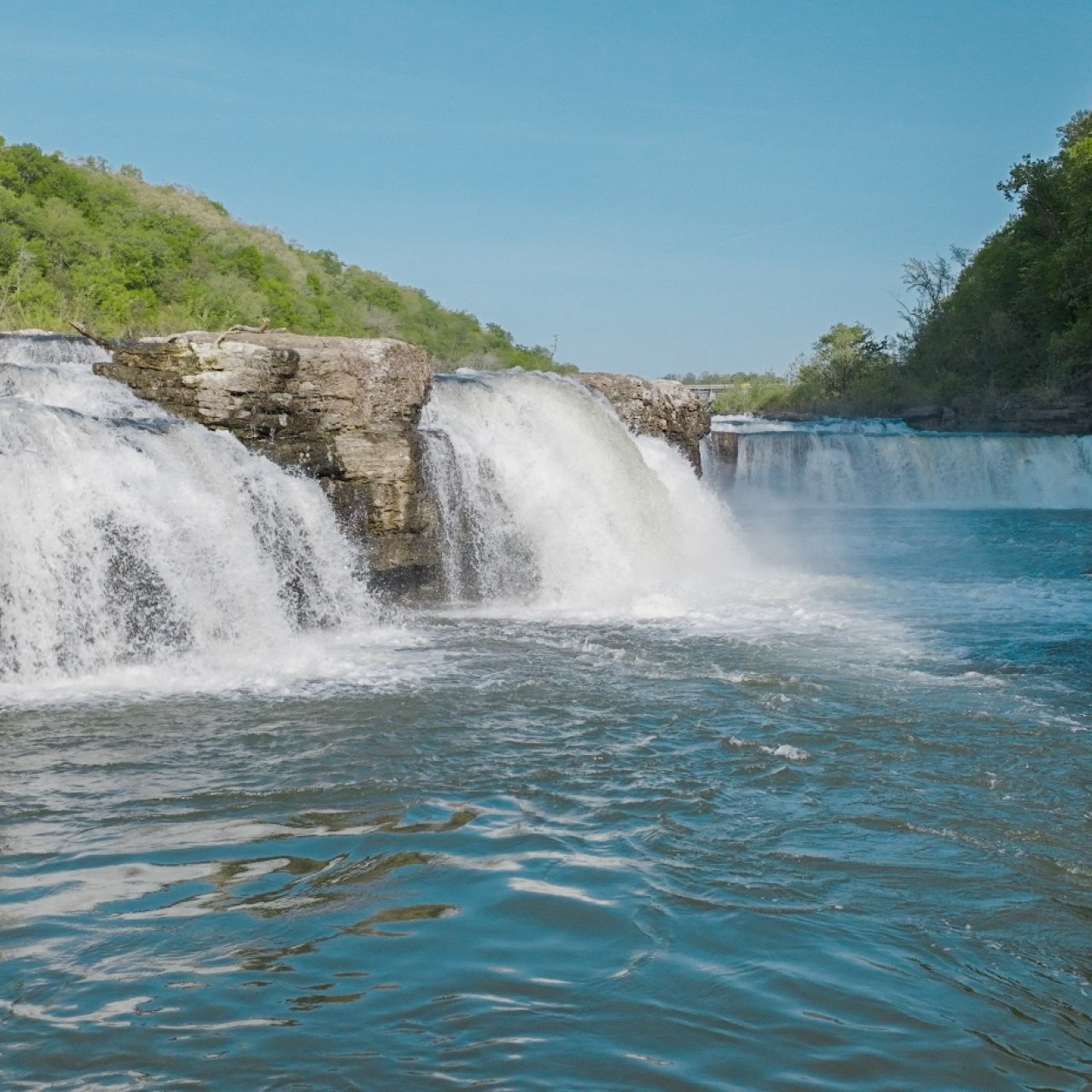 Sunny day view of a river with dual waterfalls and lush greenery.