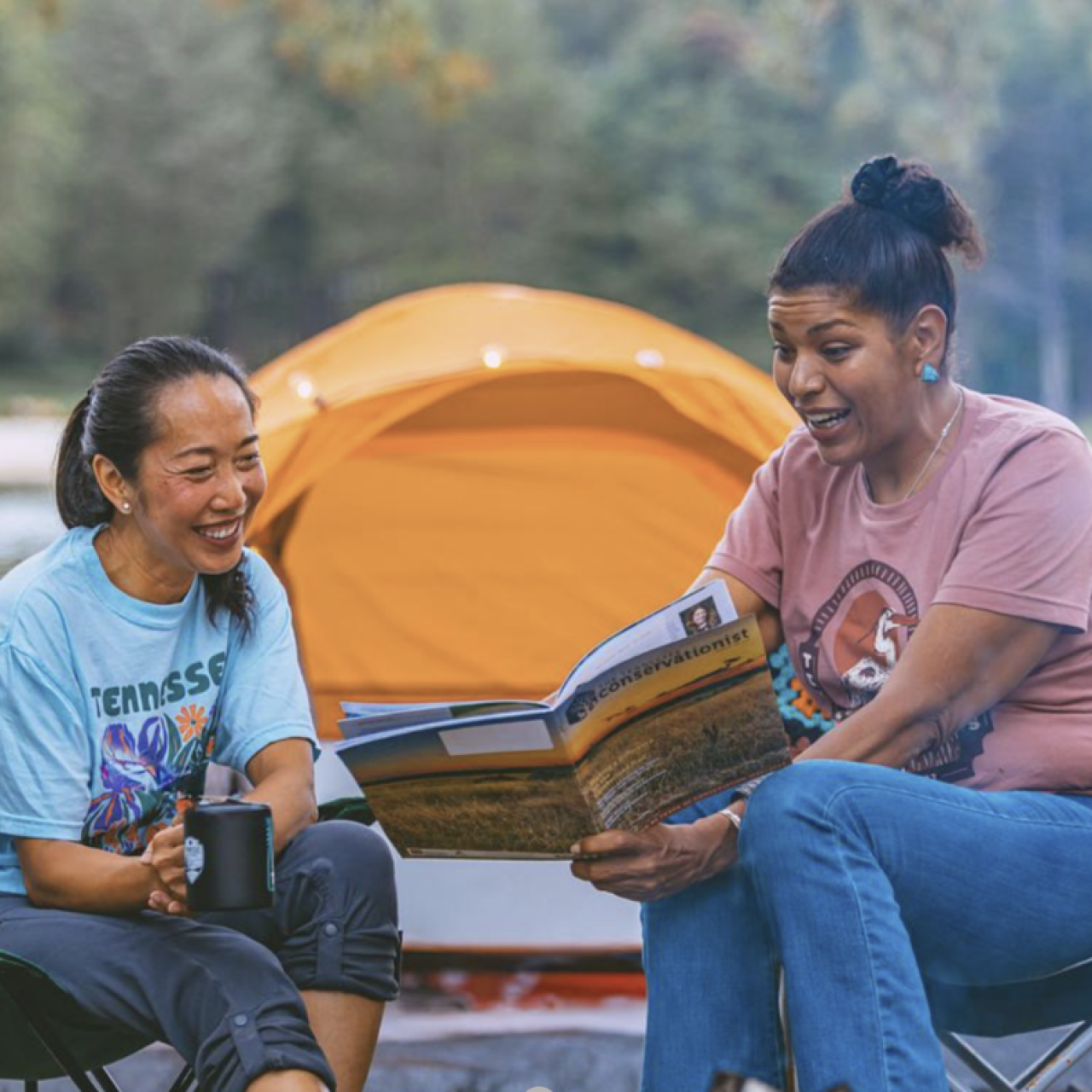 Two friends camping, reading a book near an orange tent.