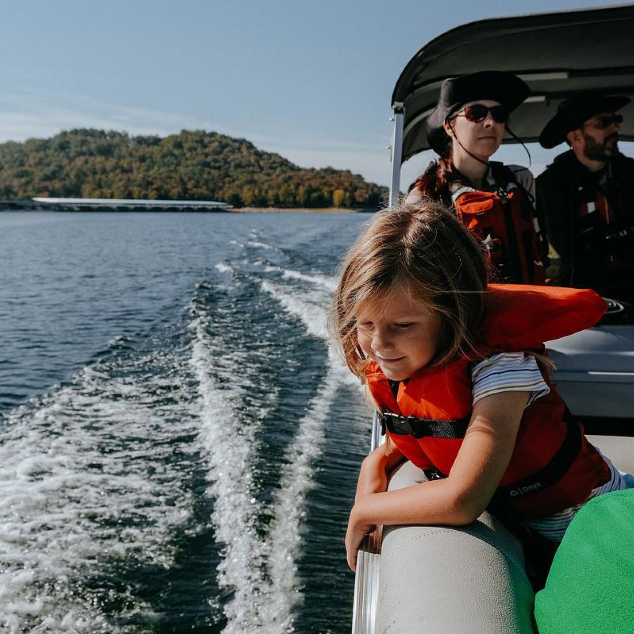 Girl in red life jacket leaning on boat railing, looking at water, with adults in background.