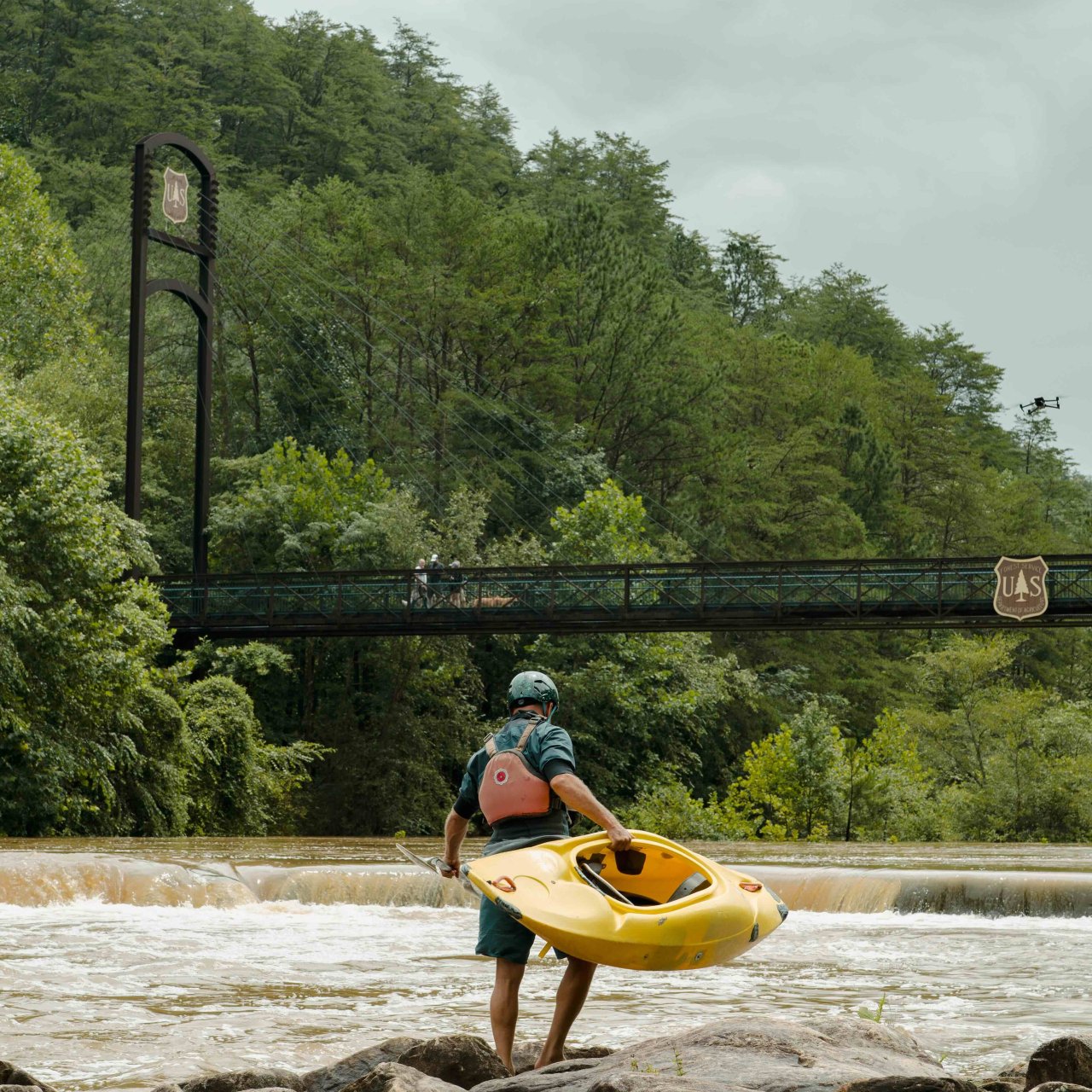 Person carrying a yellow kayak near a river with a bridge in the background.