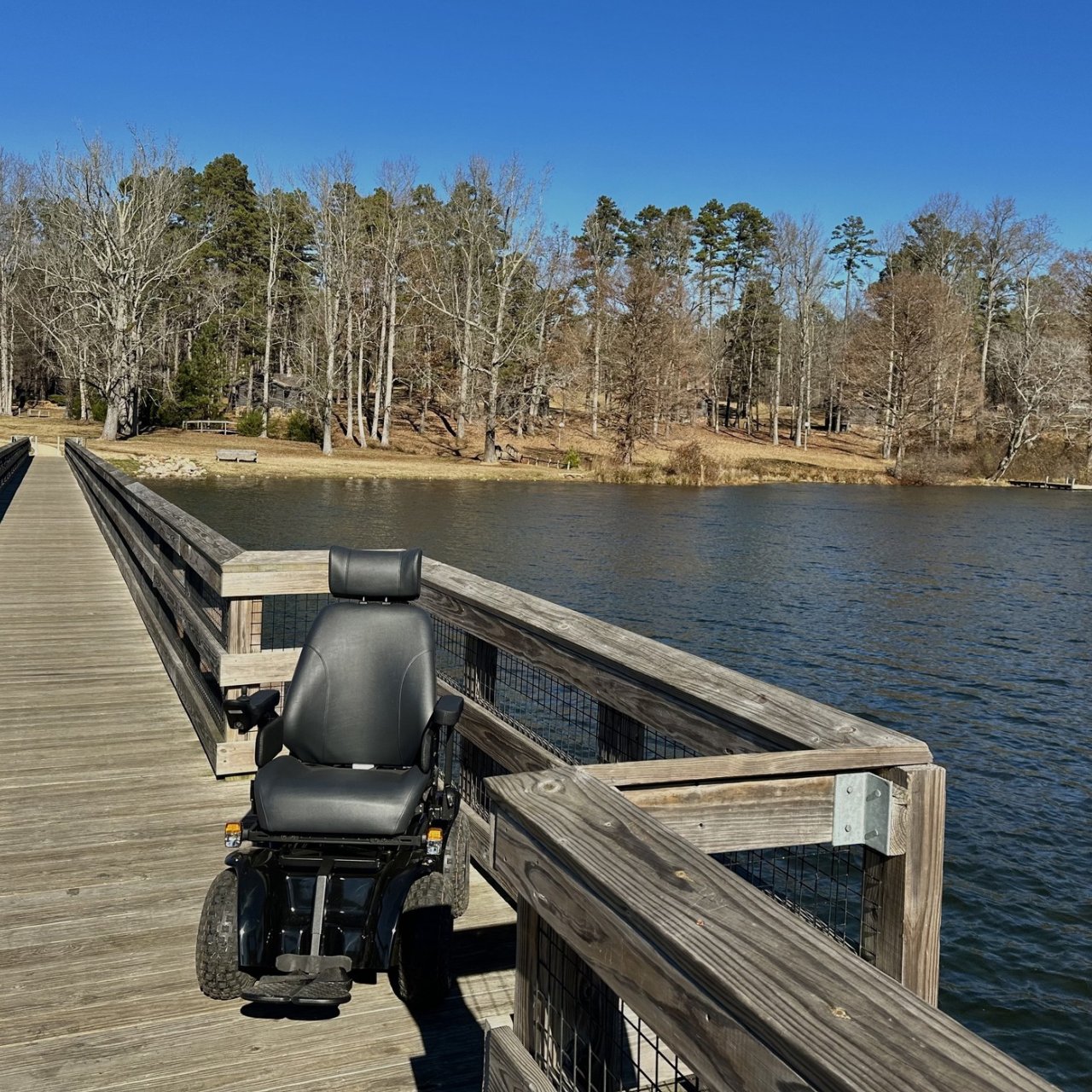 An all-terrain wheelchair sits on a boardwalk that stretches across the lake. Bare trees line the shore in the background under a bright blue sky. 