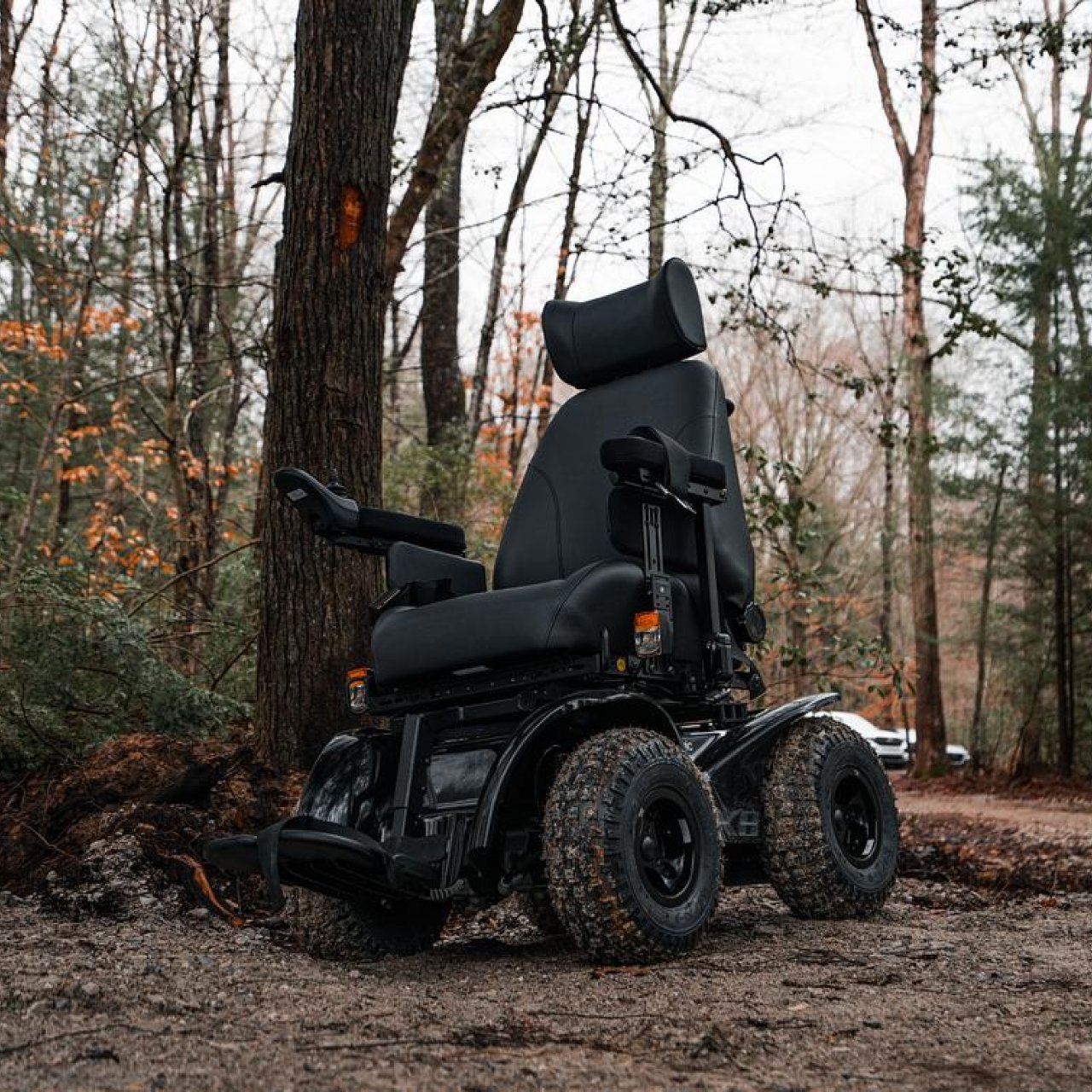 An all-terrain wheelchair sits on a natural-surfaced path. Behind the chair stand several trees that show the effects of winter on them.