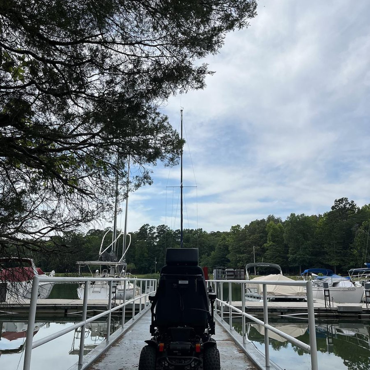 An all-terrain wheelchair sits on a bridge leading to a boat dock. Several boats are docked ahead of the chair under a cloudy sky.