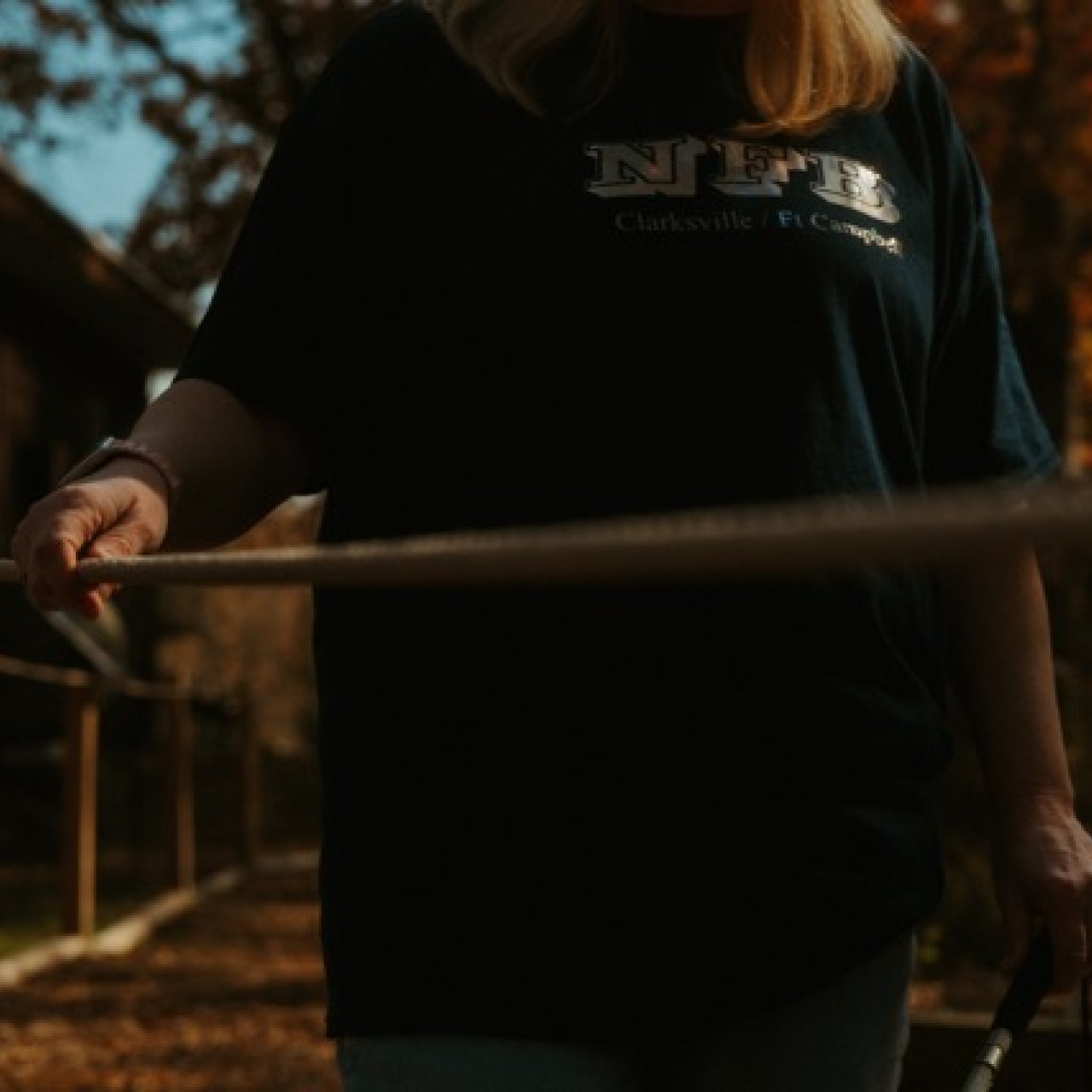 The view of a person from the shoulders down stands on a mulched path. Their right hand is placed along the guide rope as their left hand hold the handle of a white cane. In the background, there are crisp, orange leaves on the trees.