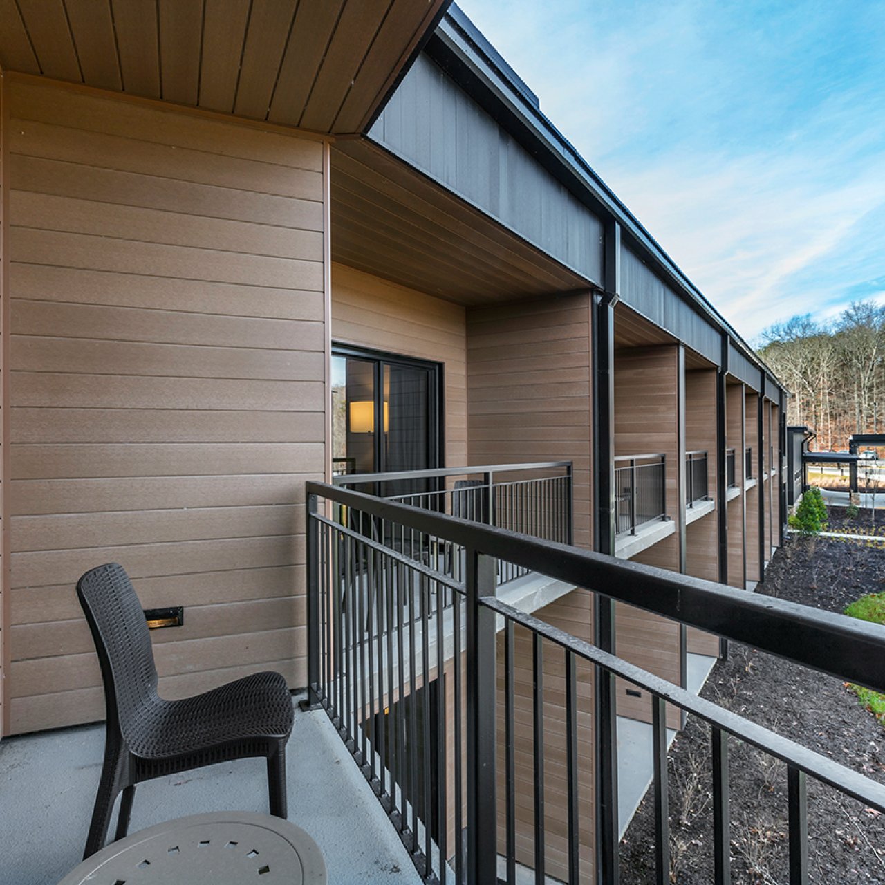 Outdoor balcony with a chair and table, overlooking a parking lot and wooded area.