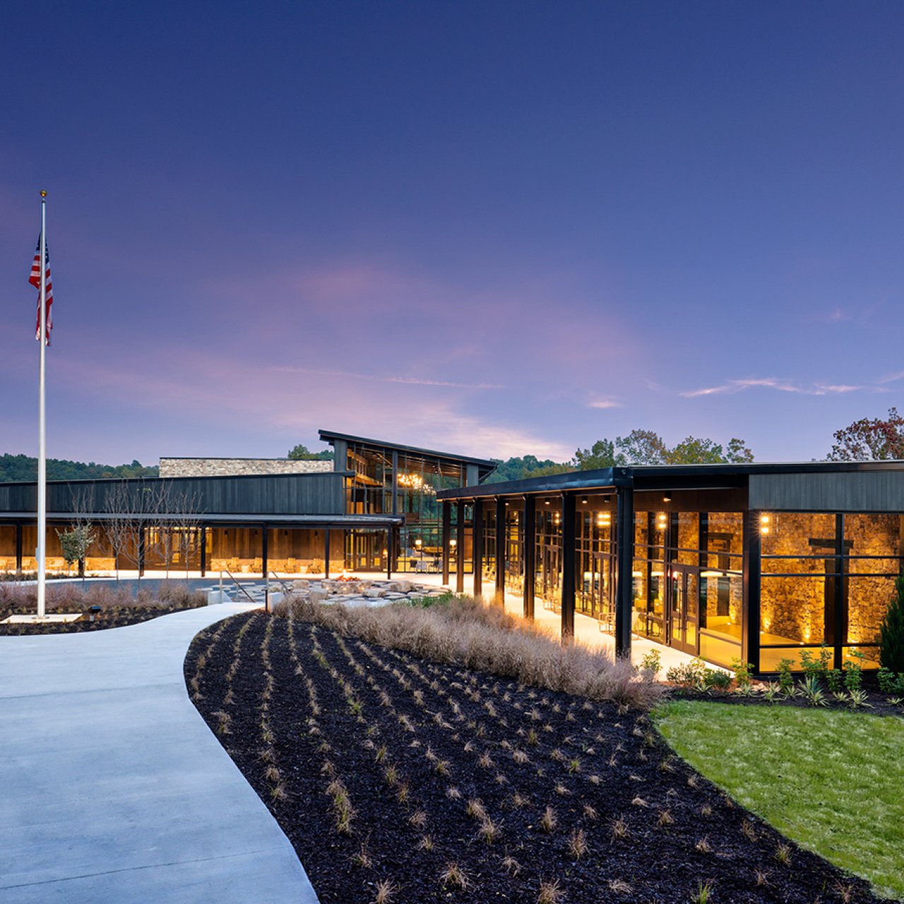 Modern building at dusk with large windows, flagpole, and landscaped path.