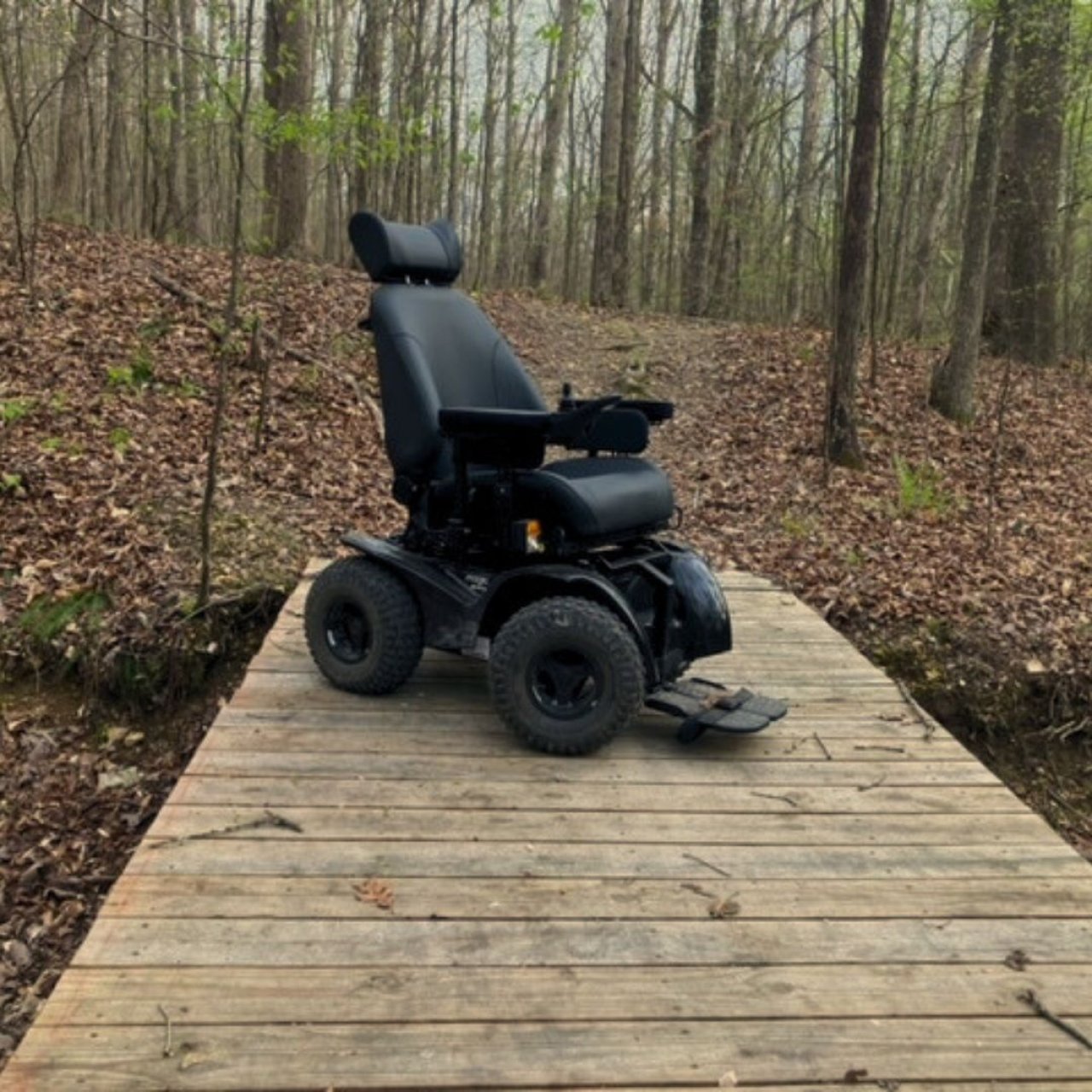 An all-terrain wheelchair sits on a boardwalk in the woods. Several trees line the background and dried leaves cover the ground.