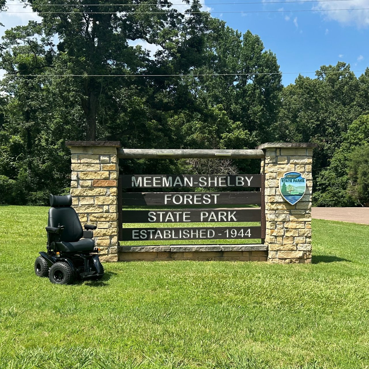 An all-terrain wheelchair sits in front of a park entrance sign that reads "Meeman-Shelby Forest State Park: Established - 1944."