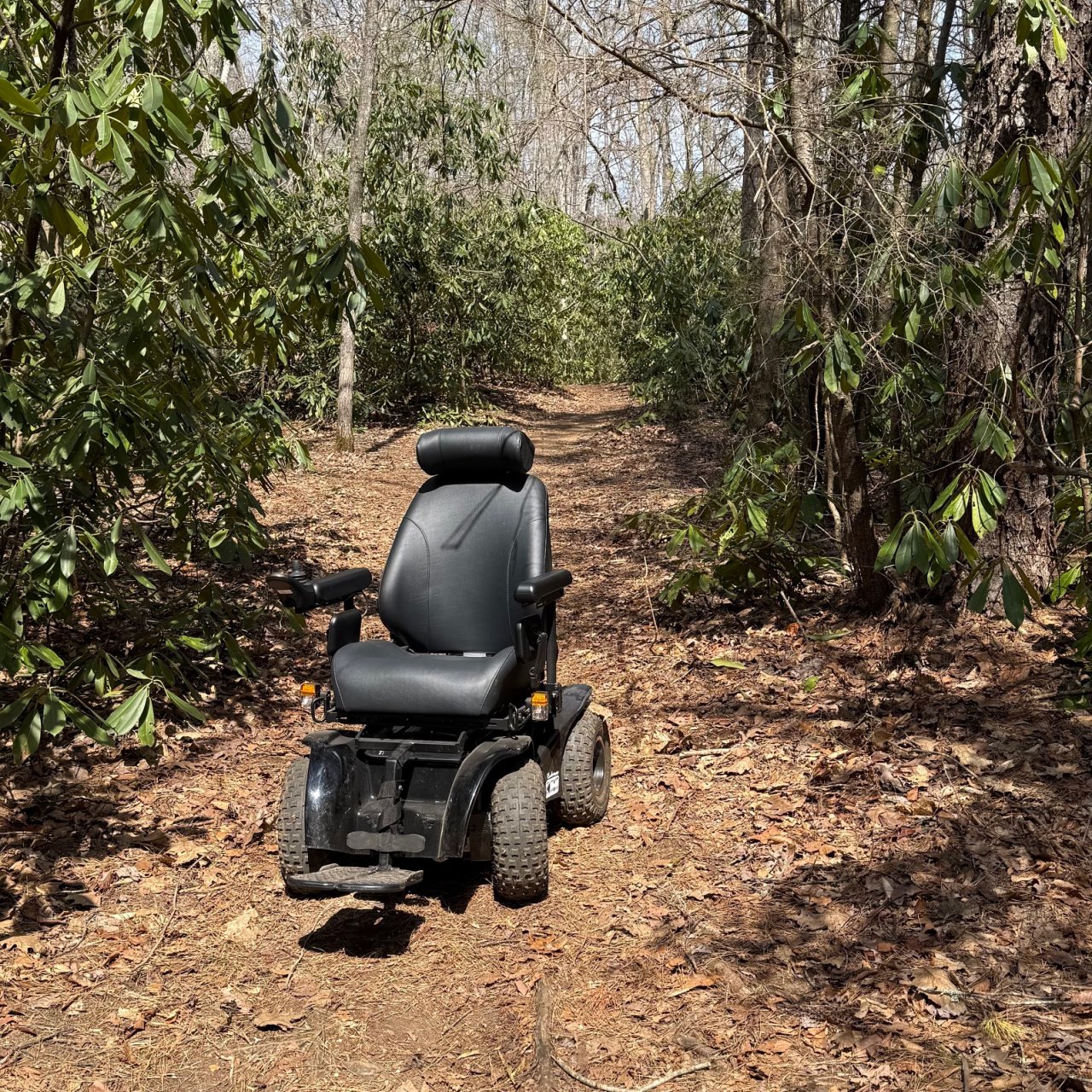 An all-terrain wheelchair sits on a natural-surfaced path that is lined with trees.