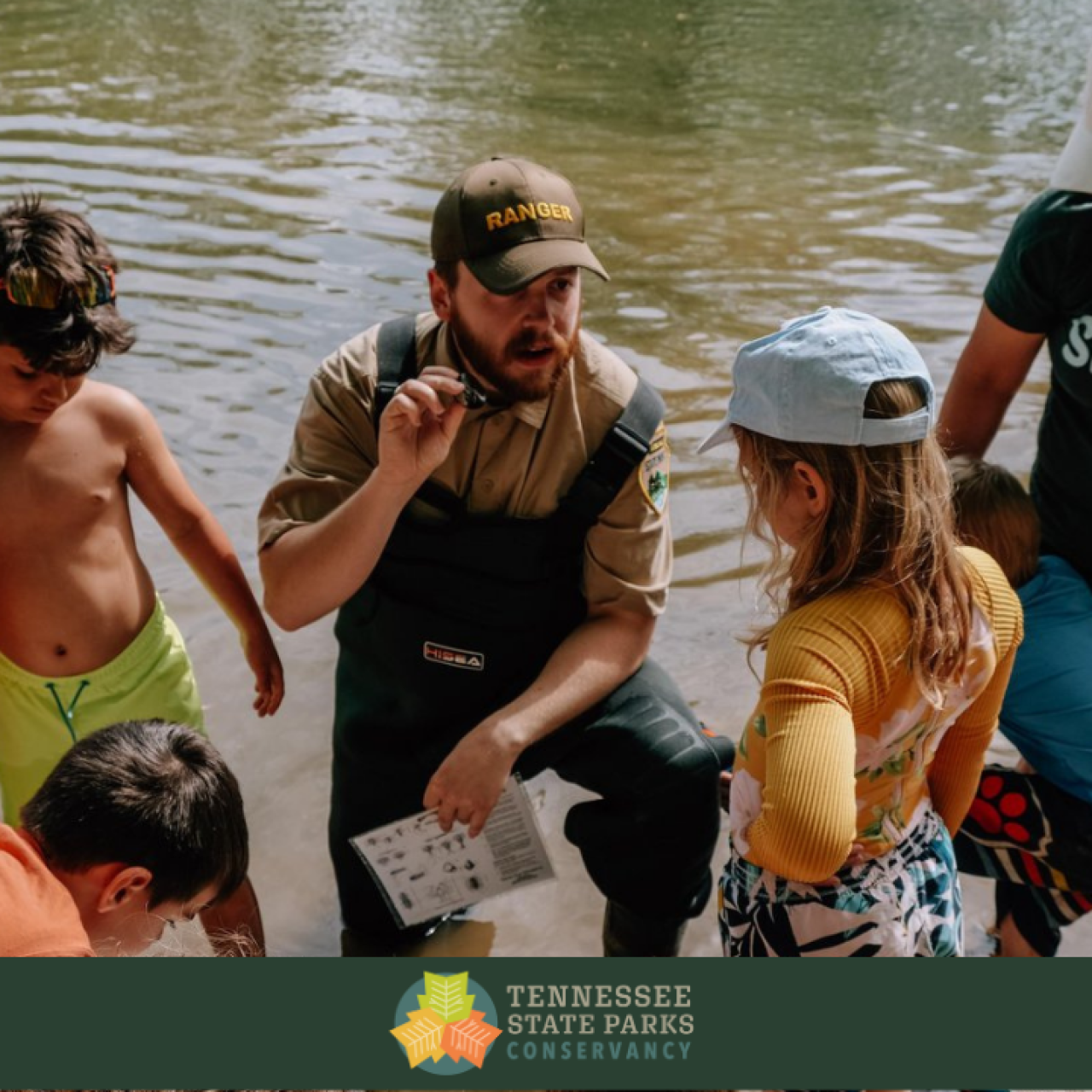 Children and a park ranger gather at a riverside for an educational moment, with everyone focused on what the ranger is explaining.