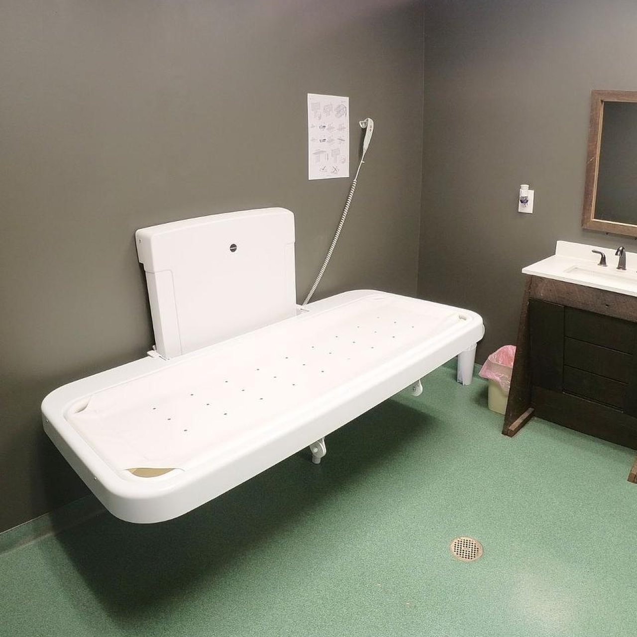 An adult-sized changing table is open in a private restroom. The restroom has gray walls and a green floor.